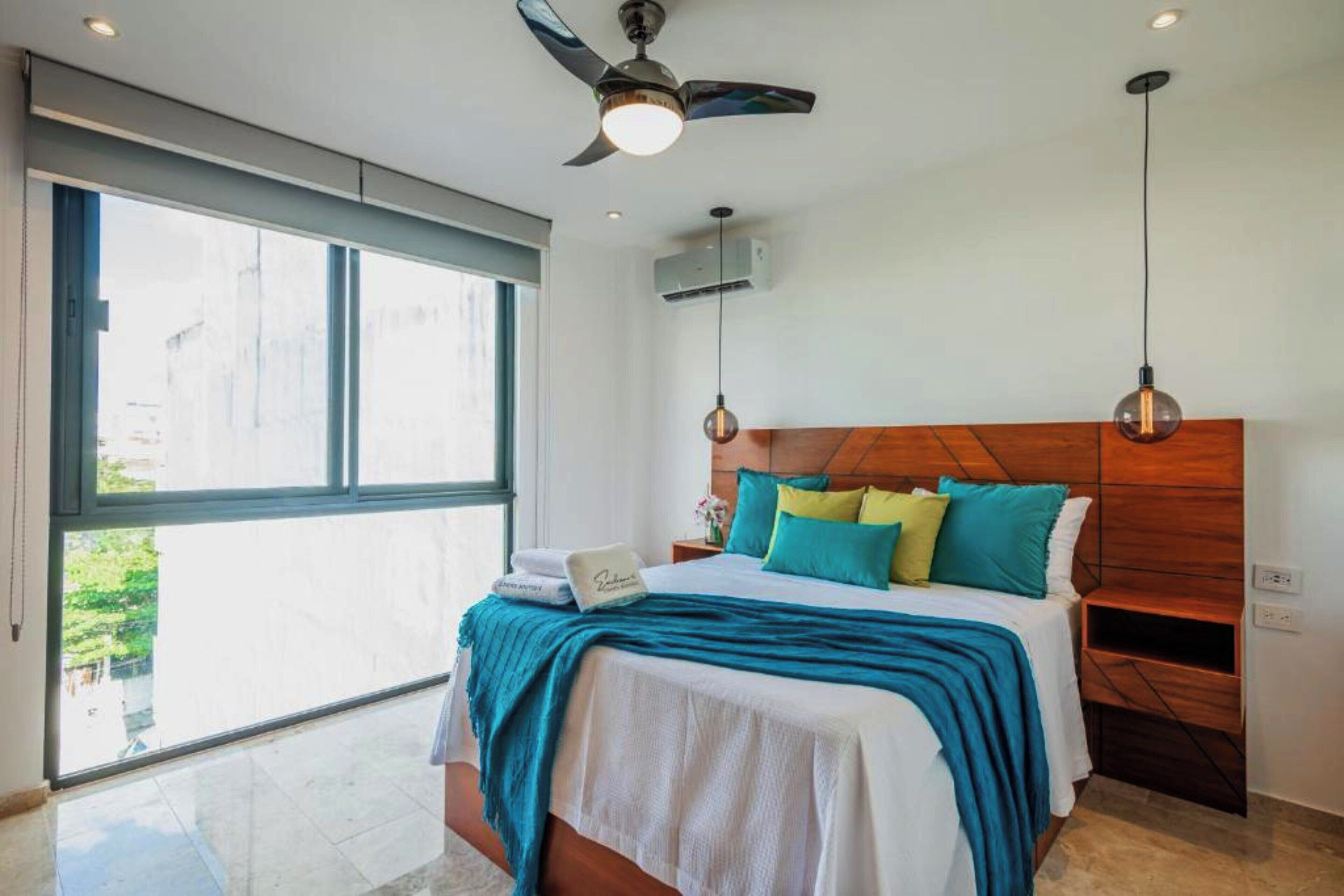 A bedroom with a large window, a ceiling fan, a bed with multiple pillows, and a nightstand.