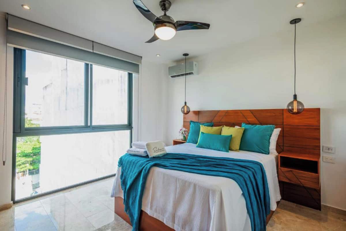 A bedroom with a large window, a ceiling fan, a bed with multiple pillows, and a nightstand.