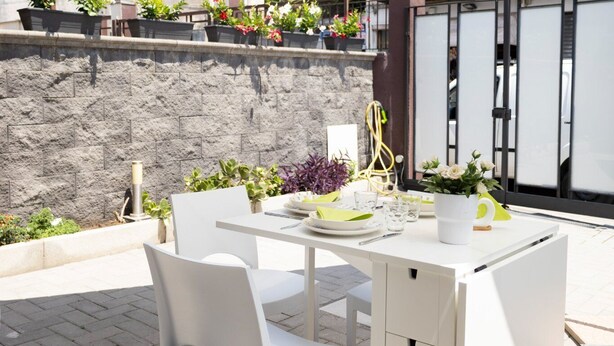Un patio al aire libre con mesa y sillas blancas, un paraguas grande y plantas en macetas.