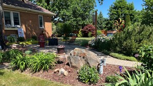 Un jardin bien entretenu, avec un grand rocher portant le chiffre « 3 406 » au centre, entouré de diverses plantes et d’un sentier menant à un bâtiment sur lequel est apposée une enseigne indiquant « The Boulder’s Bar & Grill ».