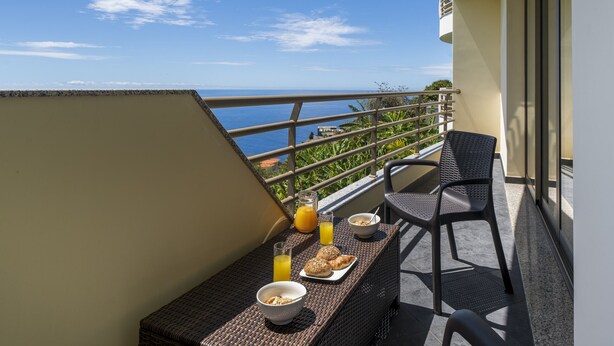 En balkon med et bord dækket til morgenmad, inklusive brød, en skål og et glas appelsinjuice.