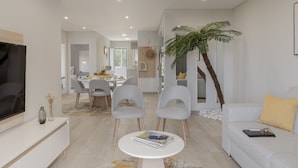 Een moderne woonkamer met een flatscreen televisie, grijze stoelen, een witte salontafel en een bank met een geel kussen.