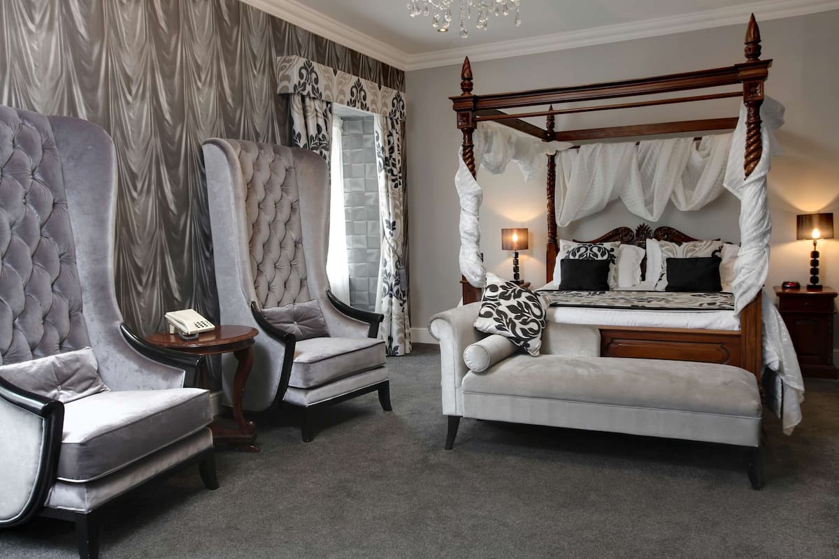 A four-poster canopy bed with a white canopy, two upholstered chairs, a small table with a telephone, and a chandelier.