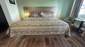Een net opgemaakt bed met een bedsprei met een patroon, een houten hoofdbord en een groene stoel naast een klein tafeltje.