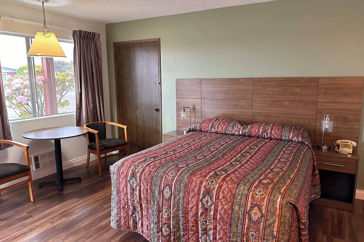 A hotel room with a large bed, a small table, two chairs, a wooden nightstand, and a lamp.