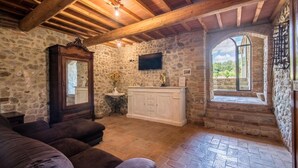 Una stanza con pareti in pietra, soffitto in legno, televisione a muro, un mobiletto bianco e una finestra con vista sul verde.