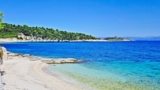 Une plage à l’eau turquoise et limpide, une rive sablonneuse et, en arrière-plan, une colline boisée.