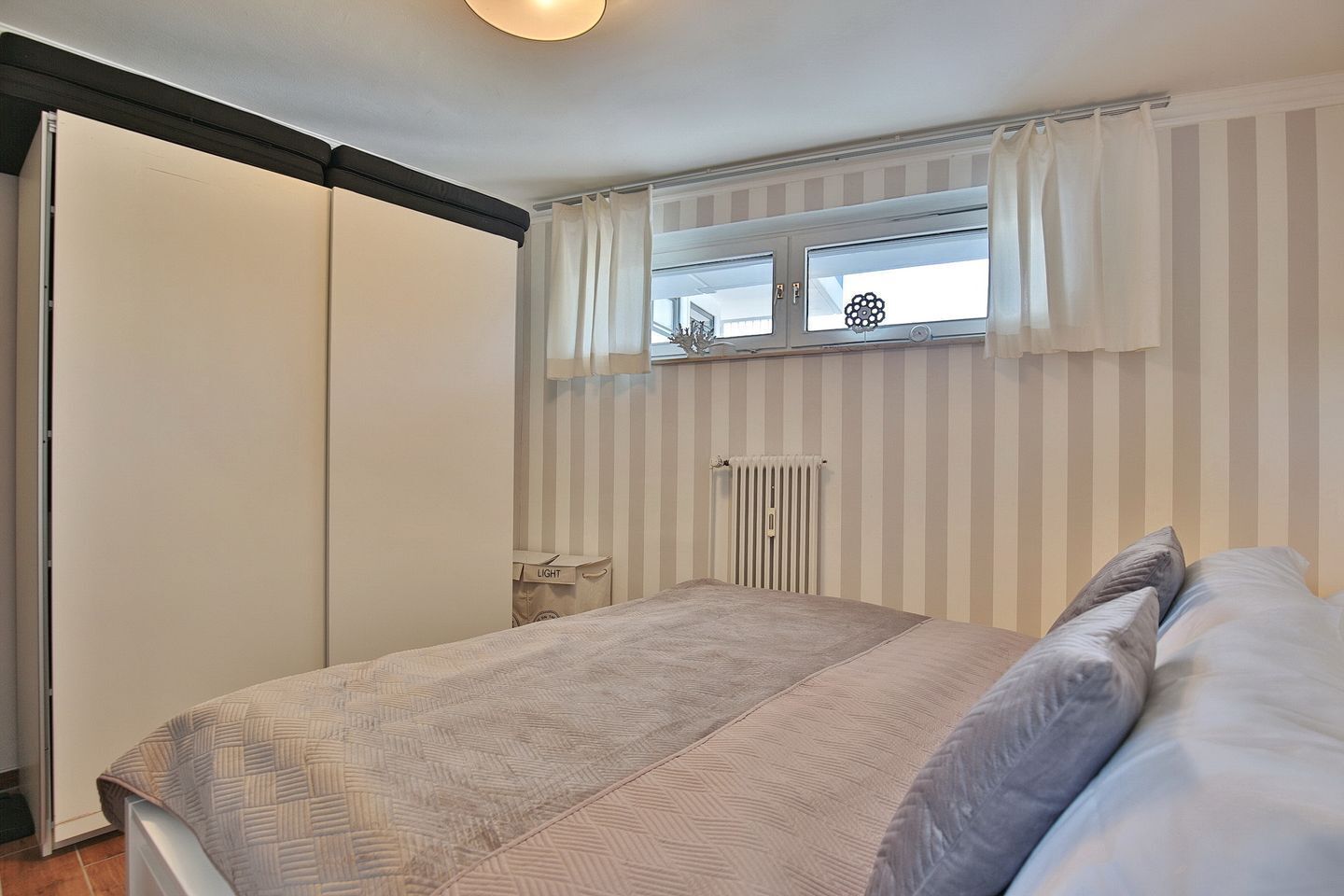 Ein Schlafzimmer mit Bett, Heizkörper, Fenster mit Vorhängen und einem Kleiderschrank.