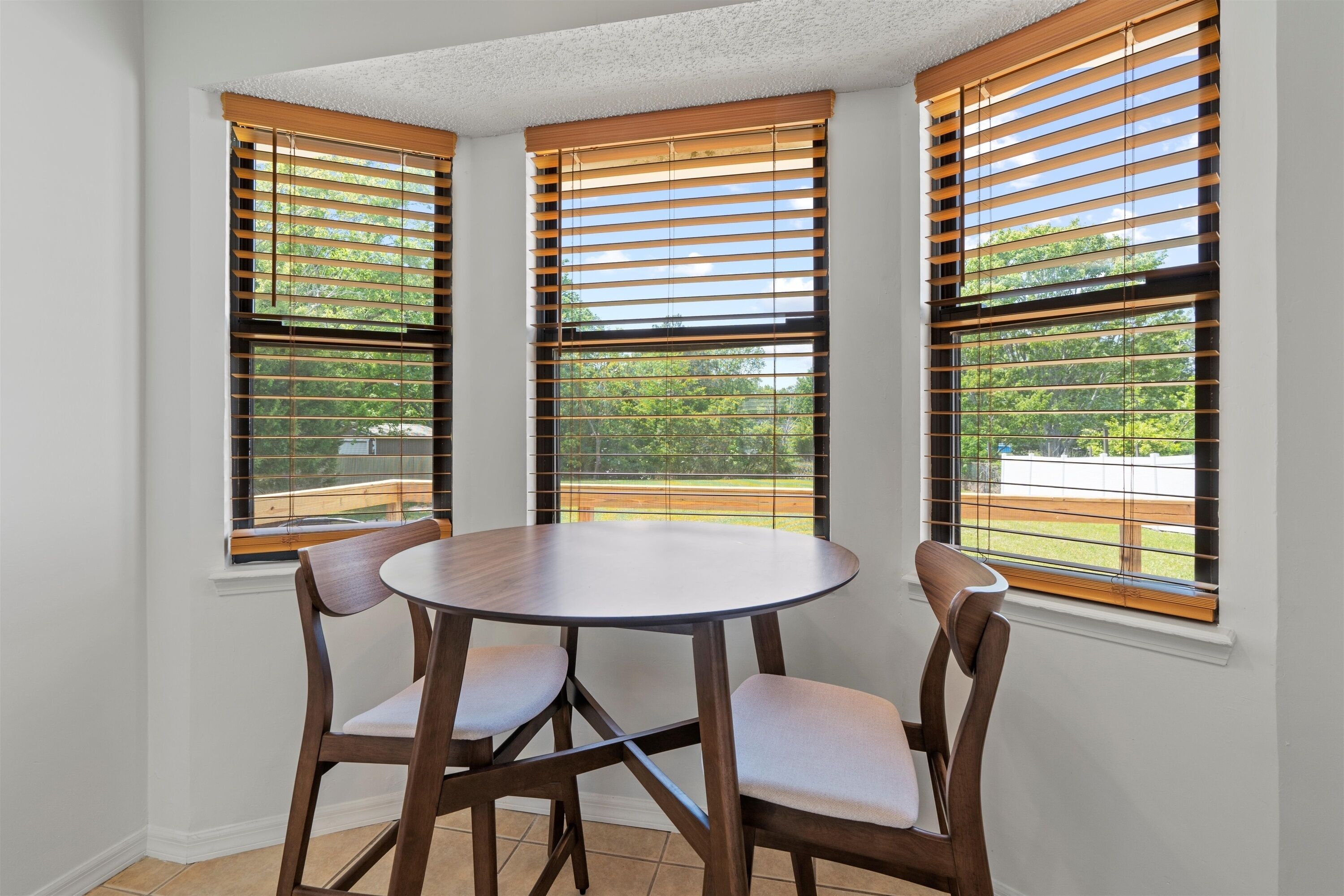 Un coin repas avec une table ronde et quatre chaises, trois fenêtres équipées de stores en bois, et une vue sur un paysage verdoyant.