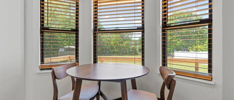 Un coin repas avec une table ronde et quatre chaises, trois fenêtres équipées de stores en bois, et une vue sur un paysage verdoyant.