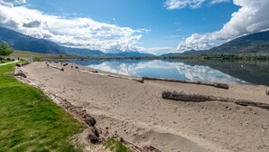 A sandy beach with logs, a calm lake, and mountains in the background. - The Lakehouse/Snow Vacation Rental (Osoyoos)