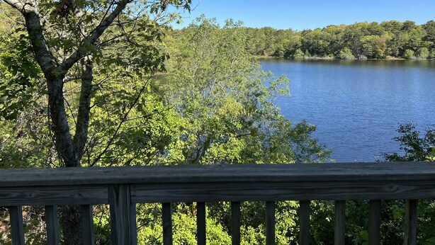 A lake surrounded by dense trees, viewed from a wooden deck. - Private Beach on Pond, Linens Included, Kayak & Canoe. All Rooms w/ Pond Views! (Chatham)