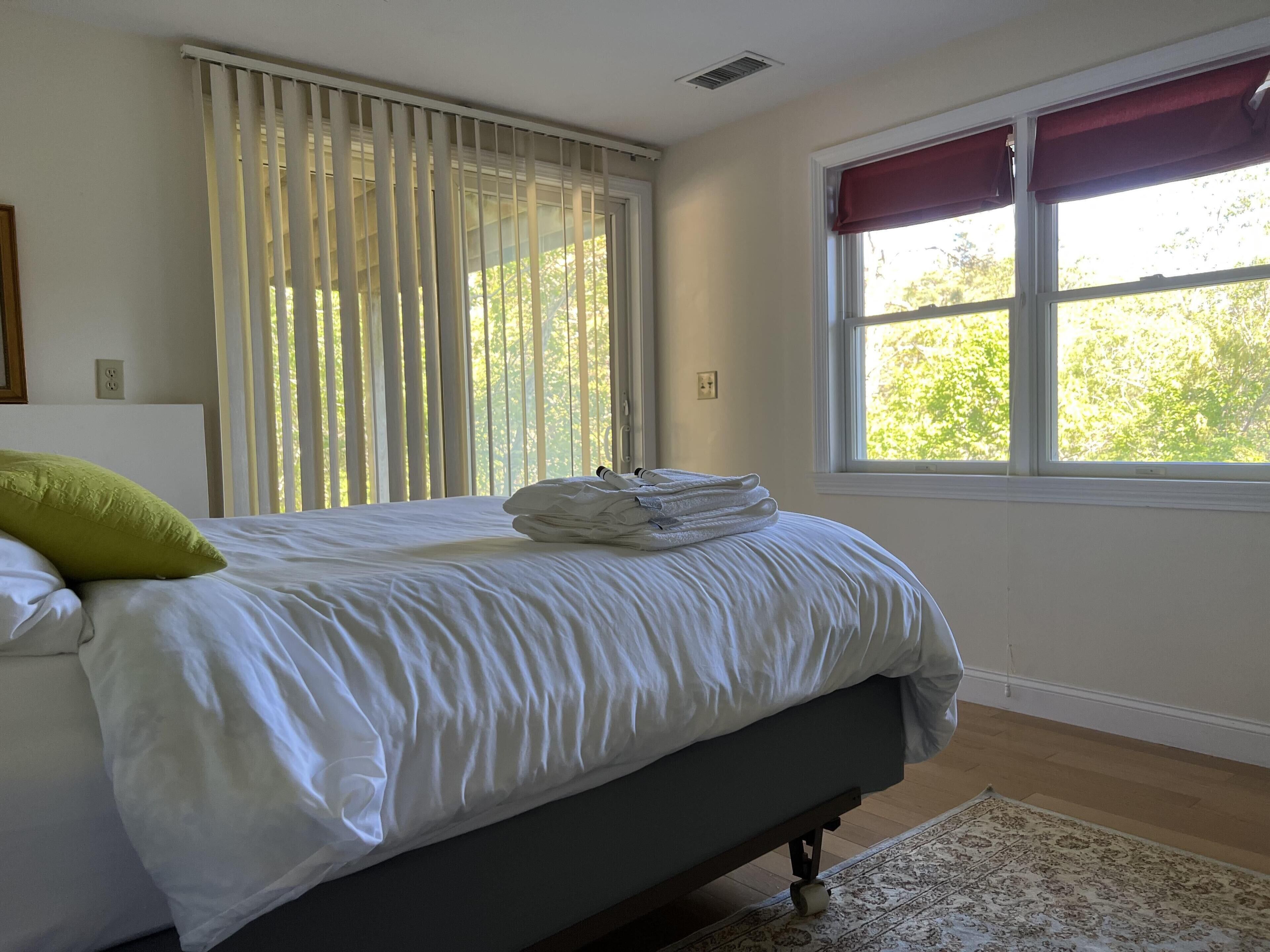 Una cama bien hecha con sábanas blancas y un almohadón verde, una ventana con un cortinero rojo, y una alfombra estampada sobre un suelo de madera.