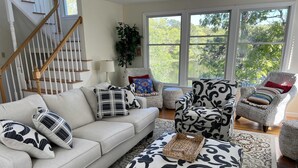 A living room with a sofa, ottoman, and armchairs, a staircase, and a large window with a view of trees. - Private Beach on Pond, Linens Included, Kayak & Canoe. All Rooms w/ Pond Views! (Chatham)