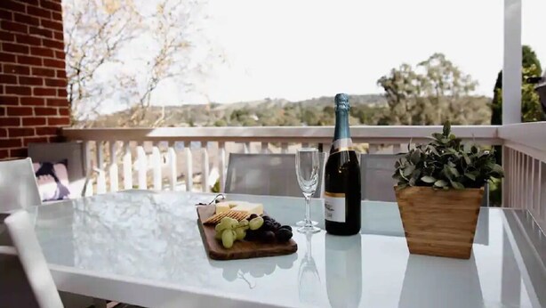 A table with a bottle of wine, a glass, and a wooden board with grapes and cheese on a balcony with a view of trees and hills. - Family, Pet  & EV Friendly Home in Bowral Centre (Bowral)