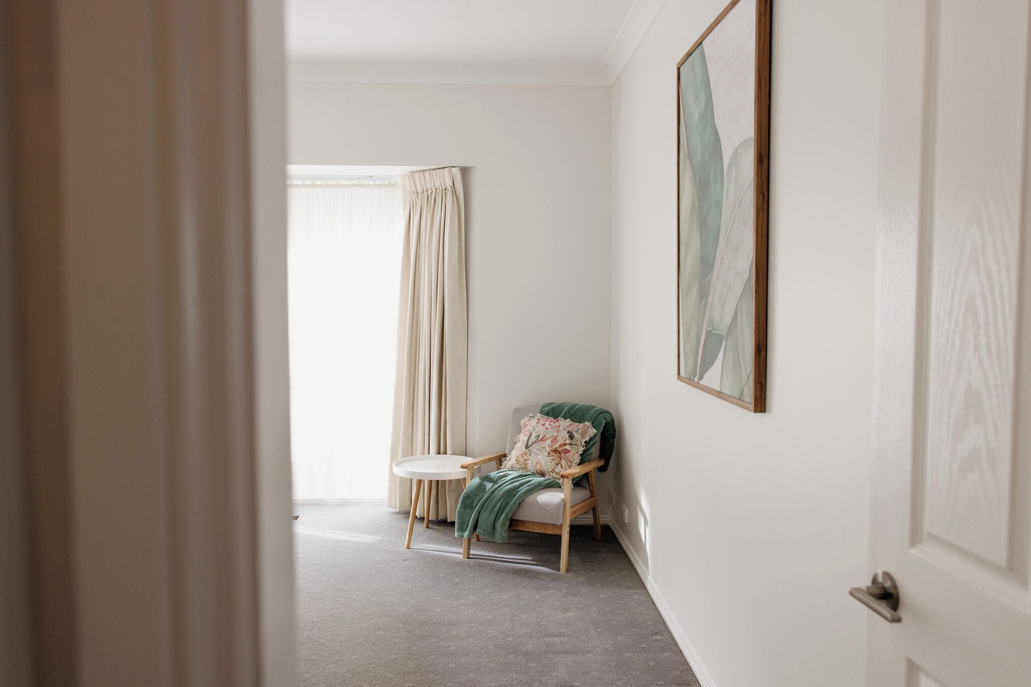 Una habitació amb una cadira, una taula rodona, una finestra amb cortines i una obra d'art emmarcada a la paret.