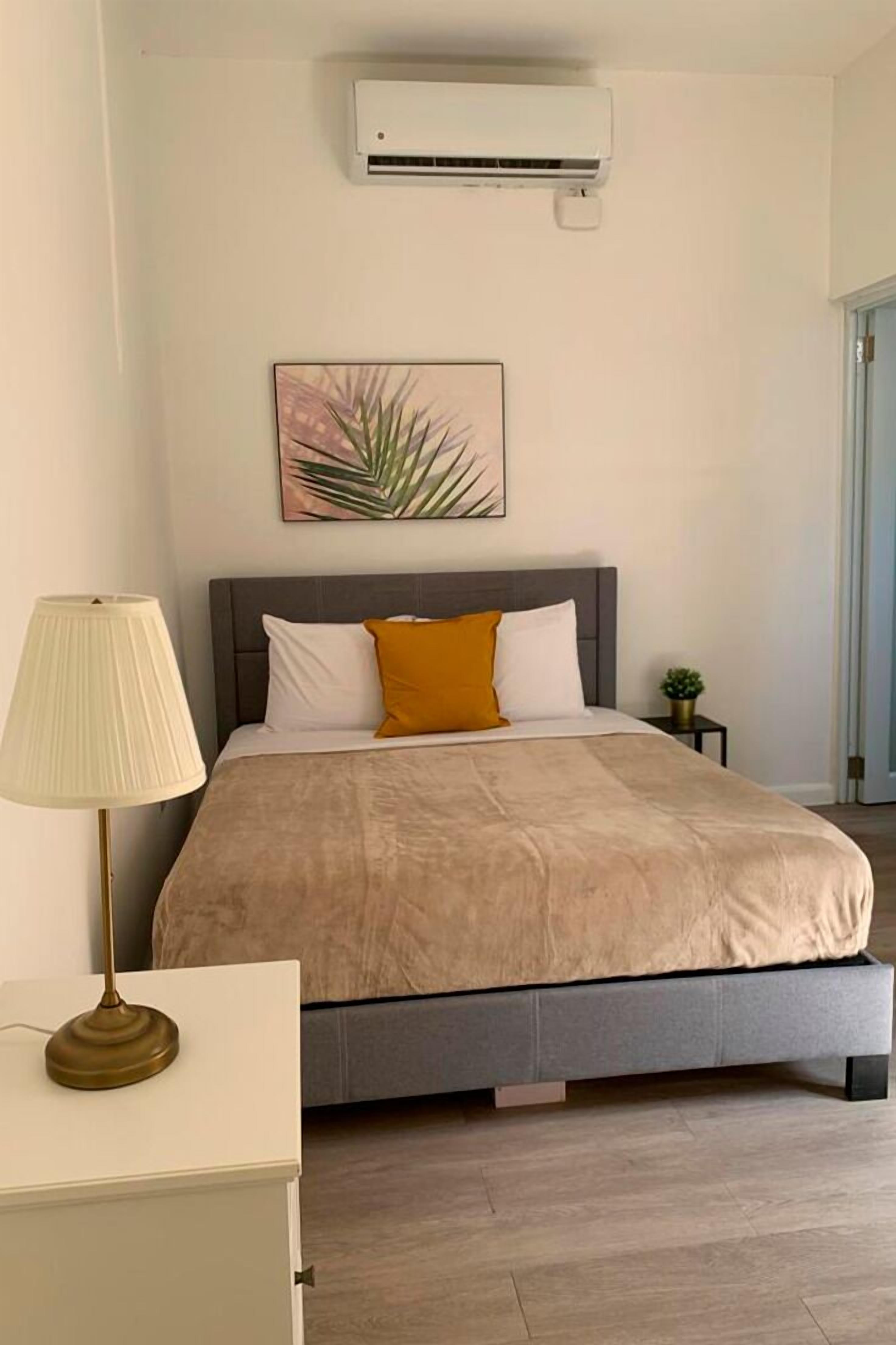 A modern bedroom with a bed, bedside table, lamp, air conditioning unit, and a small plant.