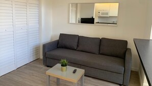 Ein Wohnzimmer mit einer grauen Couch, einem kleinen Tisch mit einer Pflanze, einer Fernbedienung und einem Blick auf eine Küche im Spiegel.