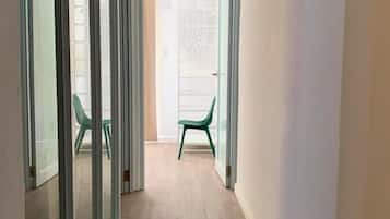 A narrow hallway with a wooden floor, glass sliding doors on the left, and a view into a room with a green chair.