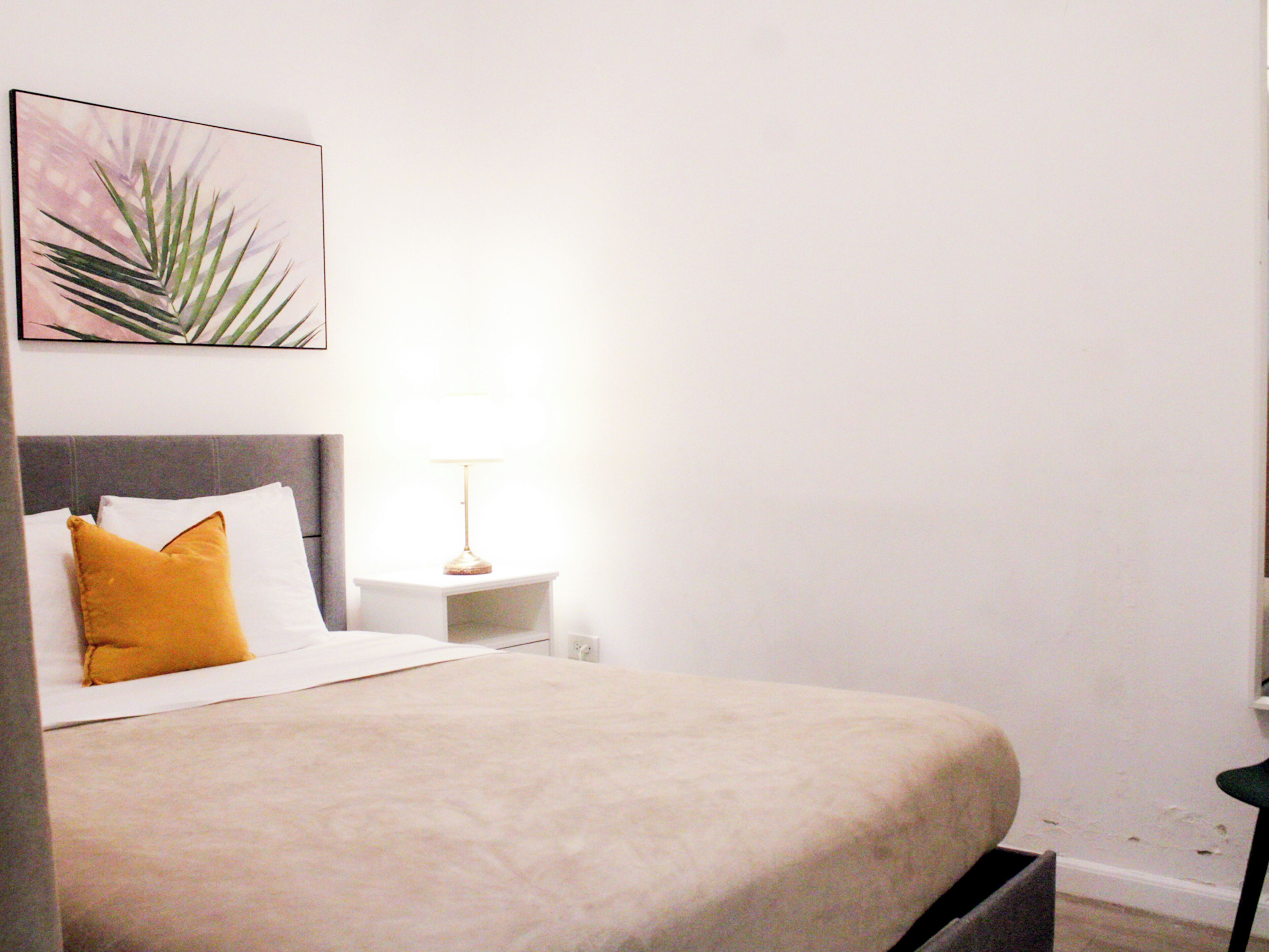 Una cama bien hecha con cubrecama color crema, almohada blanca y almohada de color mostaza. Hay una mesita de noche con una lámpara y un cuadro enmarcado en la pared sobre la cama.