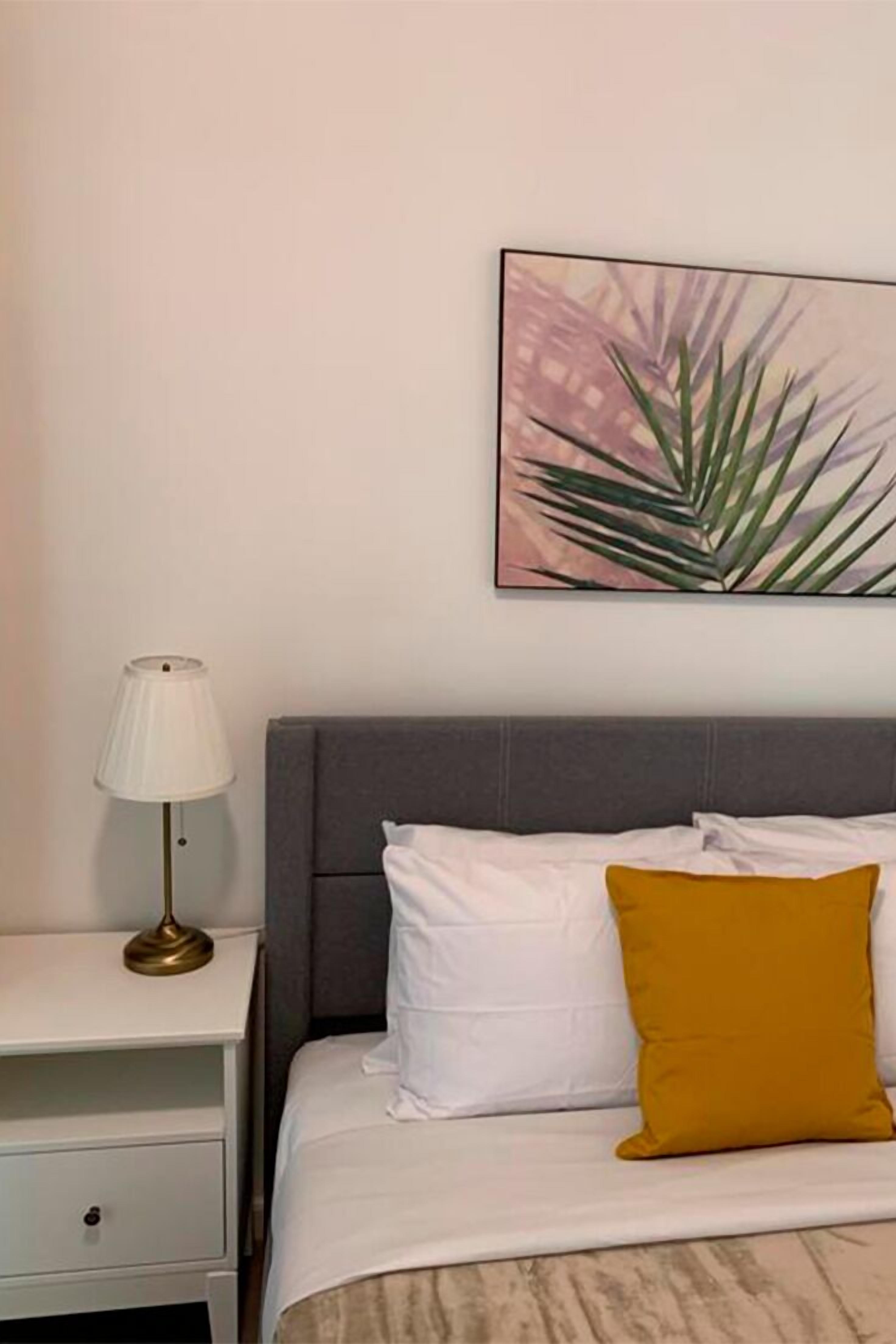 A neatly made bed with white and mustard pillows, a grey headboard, and a bedside table with a lamp.