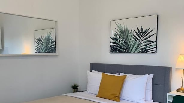 Ein ordentlich bezogenes Bett mit grauem Kopfteil, weißer Bettwäsche und einem senfgelben Kissen. Darüber an der Wand hängen zwei gerahmte Pflanzenbilder.
