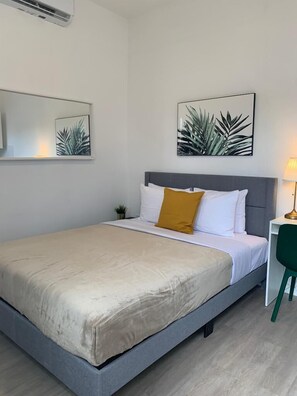 Un lit bien fait, avec une tête de lit grise, une literie blanche et un coussin de couleur moutarde. Deux reproductions encadrées de motifs botaniques sont accrochées au mur au-dessus du lit.