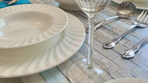 Een eettafel gedekt met witte borden, een helder glas en bestek, in een kamer met een raam en een keuken op de achtergrond.