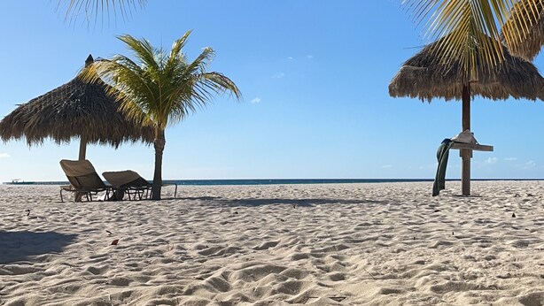 Een strand met twee rieten parasols, zand en een helderblauwe lucht.