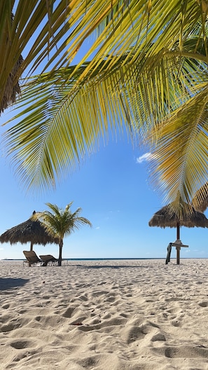 Una playa con dos sombrillas de paja, arena dorada y un cielo azul despejado.