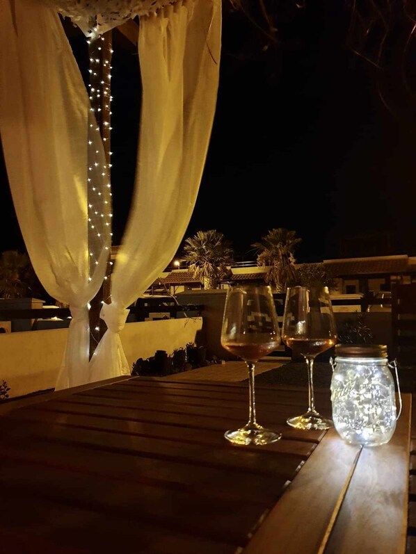 A wooden table with three wine glasses and a glass jar, set against a backdrop of draped curtains and string lights, suggesting an outdoor evening setting.
