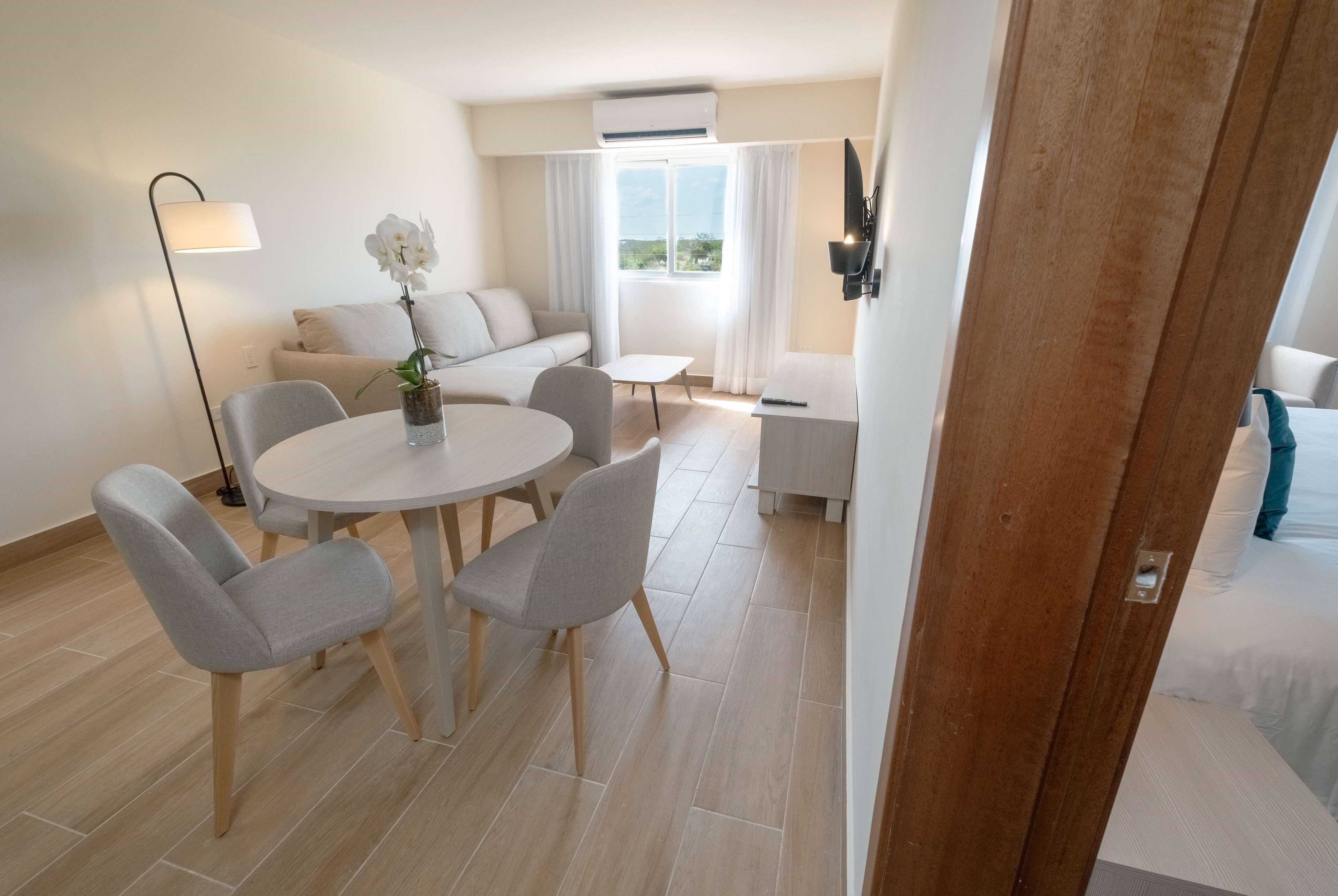 A modern hotel room with a round dining table, two grey chairs, a sofa, a small white table, a floor lamp, and a bed with a TV.