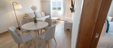 A modern hotel room with a round dining table, two grey chairs, a sofa, a small white table, a floor lamp, and a bed with a TV.