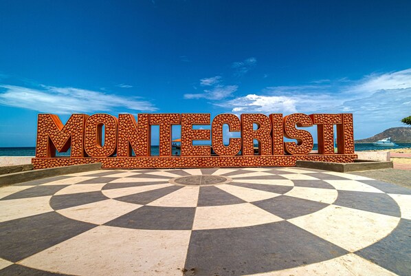 Une grande enseigne à texture marquée, sur laquelle le mot « MONTECRISTI » est inscrit en caractères gras, le tout se détachant sur un fond de ciel bleu clair et d’un pavé à motifs.