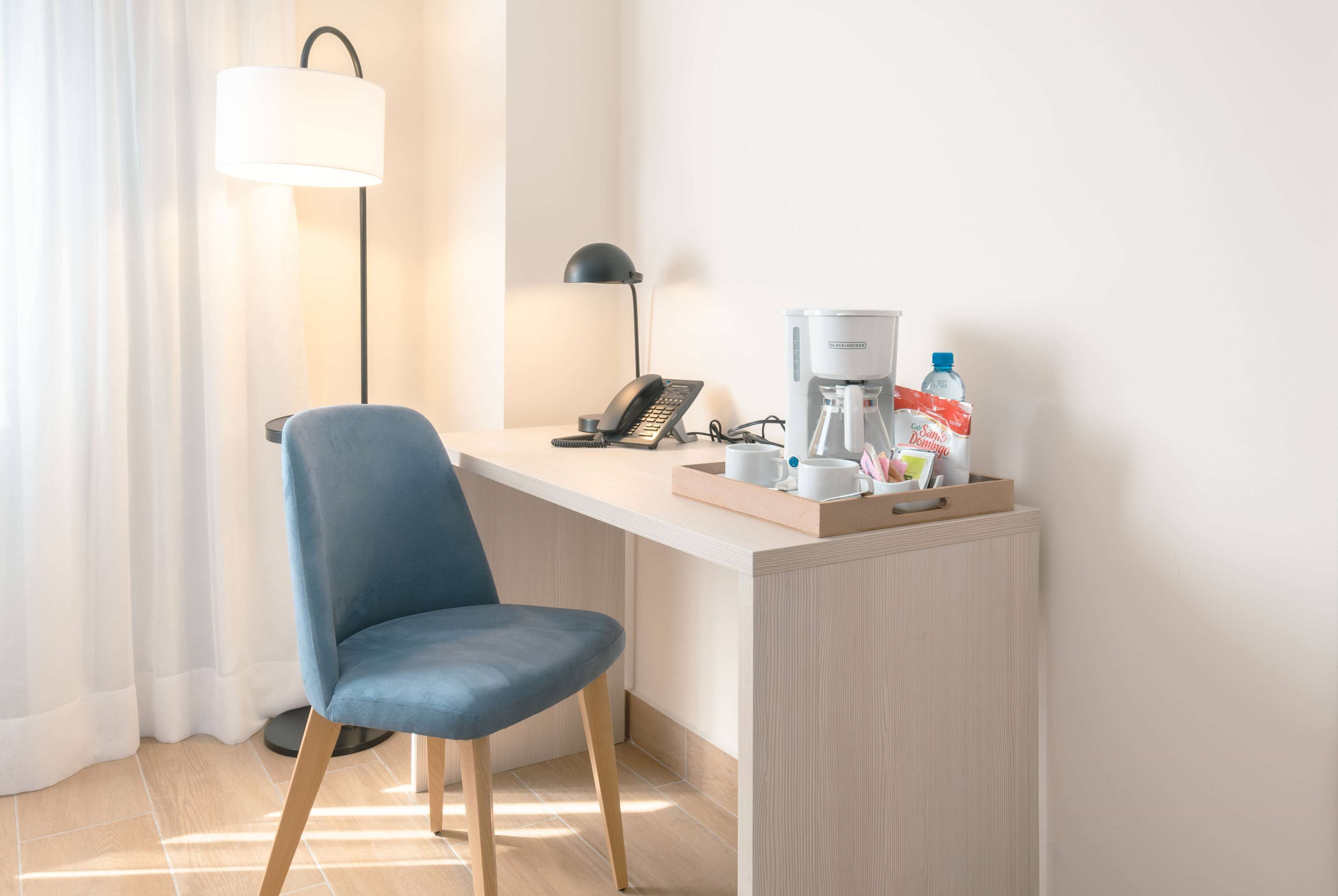 Moderni hotellihuone, jossa on työpöytä, tuoli, lamppu, puhelin, kahvinkeitin ja vesipullo.