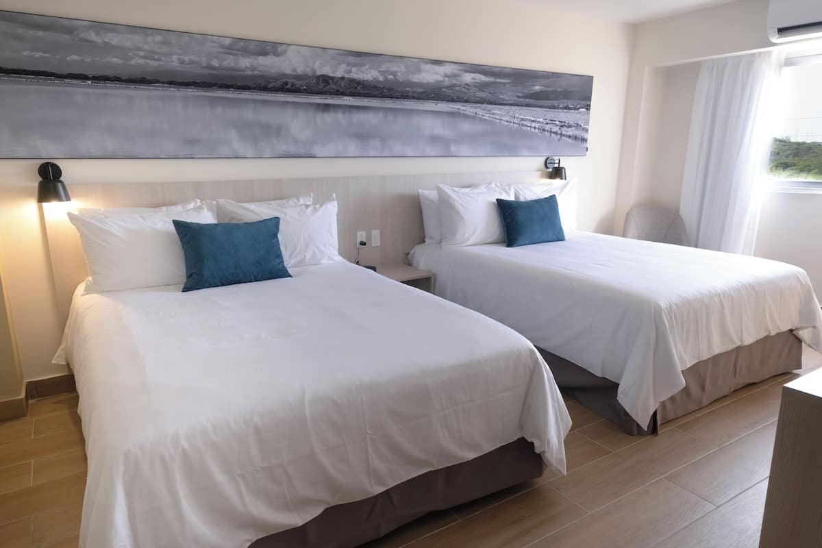 Ein Hotelzimmer mit zwei Betten, einem großen Gemälde darüber und einem Fenster mit Vorhängen.