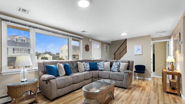 A living room with a sectional sofa, wicker coffee table, and a view of a residential area through large windows. - Vacation rental with pool on LBI (Brant Beach)