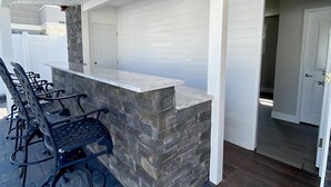 An outdoor seating area with stone-topped tables and chairs, a white brick wall, and a ceiling with recessed lighting. - Vacation rental with pool on LBI (Brant Beach)
