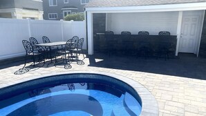 A swimming pool with a tiled edge, surrounded by a patio area with outdoor furniture and a covered seating area. - Vacation rental with pool on LBI (Brant Beach)