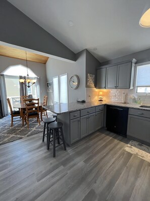 A modern kitchen with grey cabinets, a black dishwasher, and a dining area with wooden chairs and a table. - Beautiful Townhome located near the beach in Beach Haven. (Beach Haven)