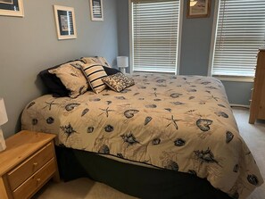A bedroom with a bed, a nightstand, a lamp, and two windows with blinds.