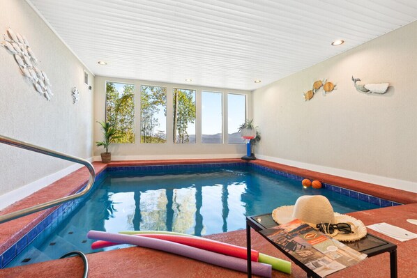 An indoor swimming pool with clear windows, a poolside table with items, and decorative wall art.