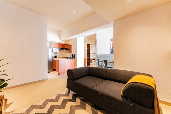 A modern living room with a sofa and a rug, leading to a kitchen with wooden cabinets and a dining area.