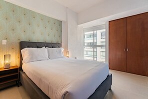A modern bedroom with a large bed, a nightstand with a lamp, a wardrobe, and a window with a view of buildings.