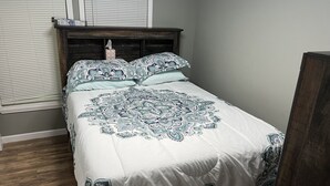 Een slaapkamer met een bed, een houten hoofdbord, een raam met luxaflex en een nachtkastje met een zakdoos.
