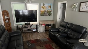 Een woonkamer met een grote flatscreen televisie, twee zwarte leren banken, een houten salontafel en een kamerbreed tapijt met dessin.