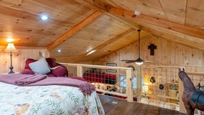 Een houten chalet met een slaapkamer op de zolder, een bed met een bloemenbedekking, een rood-wit gestreept deken, een houten plafond met zichtbare balken, een kruis aan de muur, en een paard op de achtergrond.