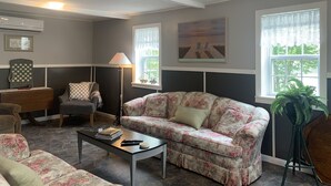 A living room with a floral sofa, a coffee table with books and a remote, a lamp, a chessboard, and a painting on the wall.