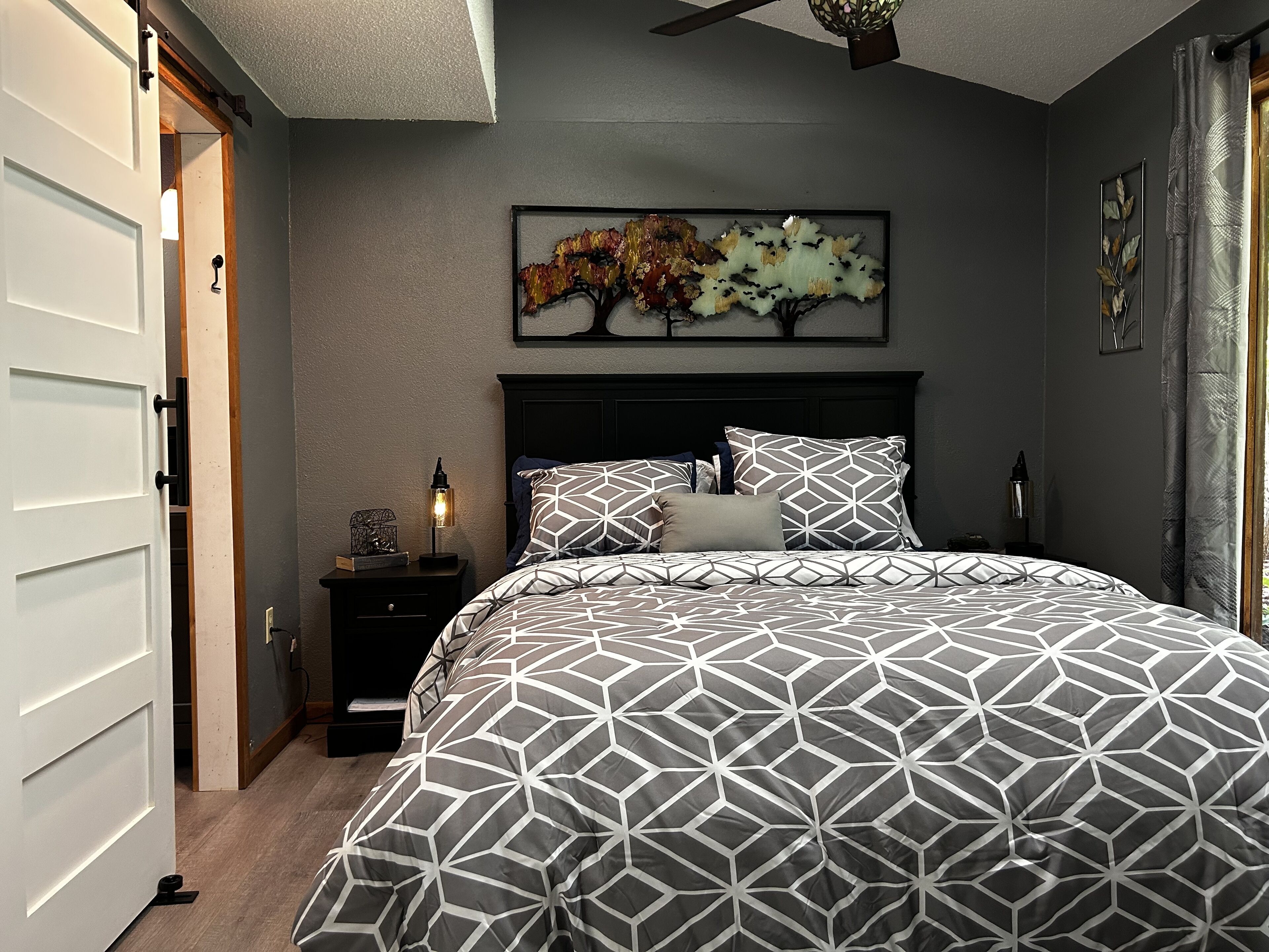 A bedroom with a large bed, a nightstand, a wall-mounted artwork, and a ceiling fan.