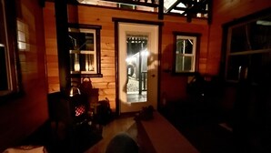 Interior de una cabaña de madera tenuemente iluminada, con una puerta que da a un balcón, una mesita con un hervidor y una silla.